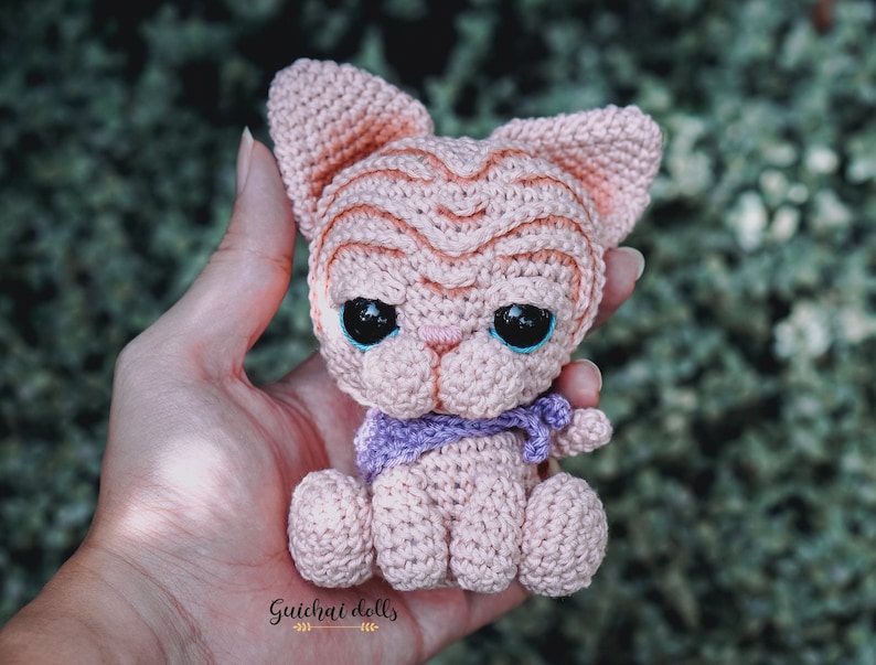 May include: A small crocheted pink cat with blue eyes and a purple scarf. The cat is sitting in a person's hand.