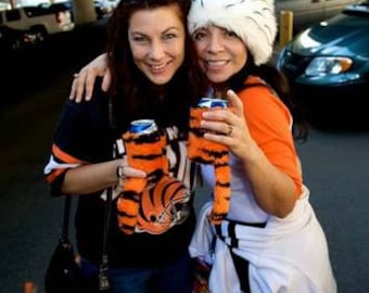 Pode incluir: Duas mulheres vestindo camisas e chapéus listrados laranja e preto sorriem e seguram latas de cerveja. Elas estão em um estacionamento.
