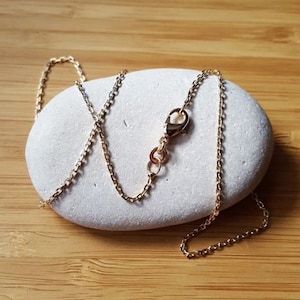Op de afbeelding: Een gouden ketting met een karabijn sluiting. De ketting ligt op een witte steen.