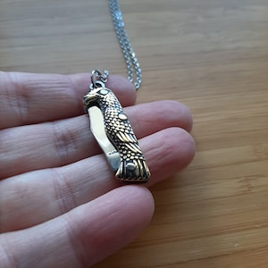 Könnte beinhalten: Ein goldfarbener Anhänger in Form eines Taschenmessers mit Adlerform an einer silbernen Kettenkette. Das Messer ist geschlossen und die Flügel des Adlers sind detailliert.