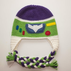 May include: A purple, green, and white crocheted hat with a felt applique of Buzz Lightyear's face. The hat has long braided ties.