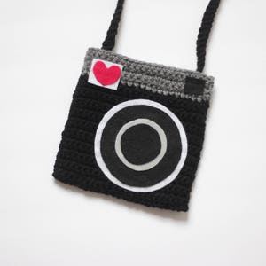 May include: A black and gray crocheted camera bag with a red felt heart on the front. The bag has a long strap and is perfect for carrying small items.