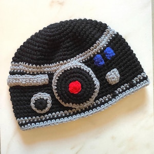May include: A handmade black crochet hat with gray and red accents. The hat features a circular design with a red center and gray trim, along with blue and gray details. The hat is designed with a unique pattern.