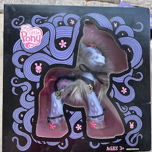 Puede incluir: Un juguete de My Little Pony en su embalaje original. La caja es negra con un diseño púrpura y el logotipo de My Little Pony. El pony es azul claro con una melena y cola grises, y está decorado con diseños florales y de llamas. La caja dice "AGES 3+".