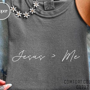 Puede incluir: Camiseta Comfort Colors gris oscuro con el texto "Jesus > Me" en fuente blanca. La camiseta es de cuello redondo y tiene una etiqueta que dice "Comfort Colors". Un collar plateado con colgantes de flores está sobre la camiseta.