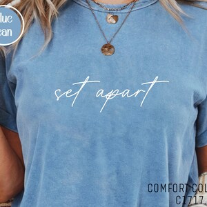 May include: Blue jean Comfort Colors C1717 t-shirt with the words "set apart" in white script. The shirt has short sleeves and a crew neck. The shirt is a light blue color.