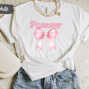 May include: White t-shirt with the word "Passover" in pink script above a pink bow graphic. The shirt is styled with blue jeans and a cream-colored purse. A gold necklace is also visible.