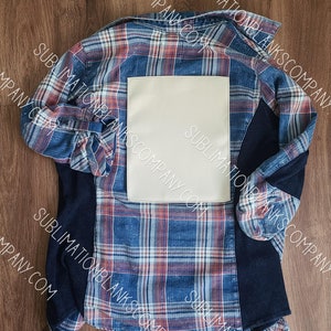May include: A blue and red plaid flannel shirt with a blank white patch on the back. The shirt has a navy blue sleeve and is laid flat on a wooden surface.
