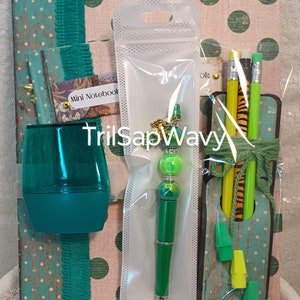 May include: A green and gold pen with a green and white bead charm, a green pencil holder with three pencils, a green and white mini notebook, and a green plastic pencil sharpener. The items are arranged on a pink and green polka dot background.
