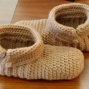 May include: A pair of crocheted slippers in a light brown colour. The slippers have a button detail on the top of the foot.