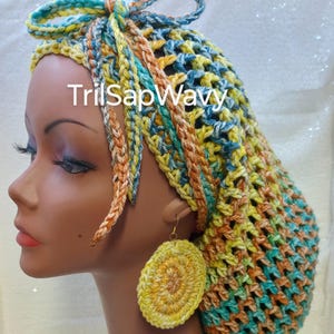 Op de afbeelding: Een gehaakte haarnetje in tinten teal, geel en oranje met een strik bovenop. Het haarnetje wordt gedragen door een mannequin met bijpassende gehaakte oorbellen.