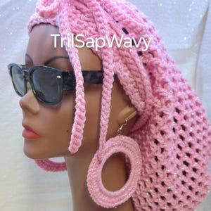 May include: Pink crochet hat with long braids and large pink hoop earrings. The hat has a sparkly texture. The text "TrilSapWavy" is visible on the hat.