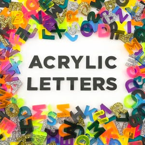 An assortment of various colorful acrylic letters scattered around the edges to create a frame around the words Acrylic Letters that is cut out of dark grey acrylic.