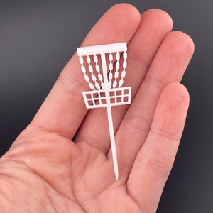 A single white miniature disc golf basket cupcake topper held in a hand to show scale.