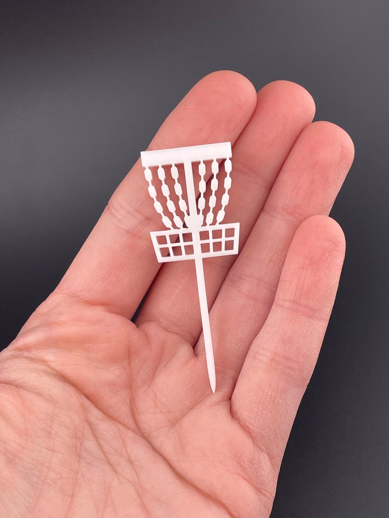 A single white miniature disc golf basket cupcake topper held in a hand to show scale.