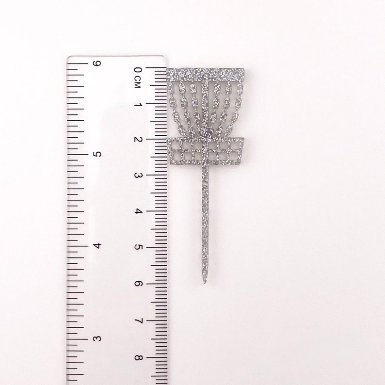 A single silver glitter acrylic disc golf basket cupcake topper against a ruler to show the height of 6 centimeters tall.