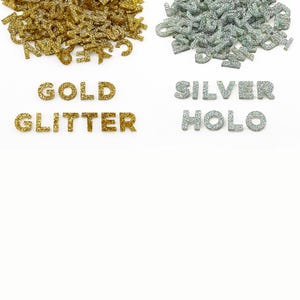 Two piles of half inch capital letters cut from acrylic. One pile of metallic gold glitter letters and another pile of holographic silver glitter letters that shine rainbow colors when the light reflects off the glitter.