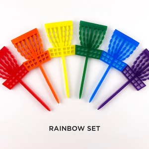 May include: A set of six rainbow-colored disc golf discs with a unique design. The discs are arranged in a semi-circle with the colors red, orange, yellow, green, blue, and purple. The text "RAINBOW SET" is printed below the discs.