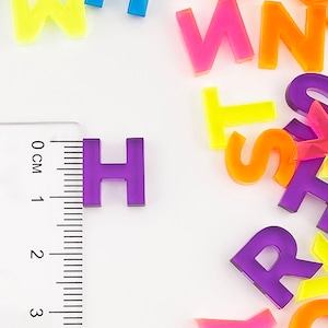 May include: A close-up of a purple letter H made of plastic. The letter is sitting on a white surface next to a ruler. The ruler is marked in centimeters.