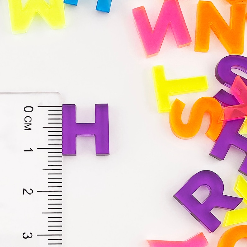 May include: A close-up of a purple letter H made of plastic. The letter is sitting on a white surface next to a ruler. The ruler is marked in centimeters.