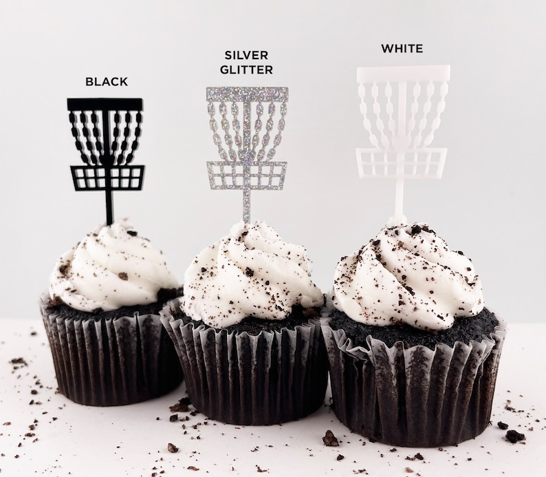 3 mini chocolate cupcakes with vanilla icing and cookie crumbles, decorated with tiny disc golf basket cupcake toppers in black, silver glitter, and white.
