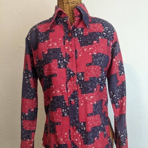 Op de afbeelding: Een button-down shirt met lange mouwen en een patchwork-ontwerp. De shirt heeft een patroon van rode, marineblauwe en witte bloemen en geometrische vormen. Het heeft een puntkraag en rode knopen.