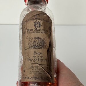 May include: A clear glass bottle with a red and blue cap. The bottle has a label with the text "MAD MADUSA'S TREACHEROUS ODDITIES" and "SPAWN of Ia O'Lanier". The bottle contains an orange liquid, and is likely a Halloween-themed item.