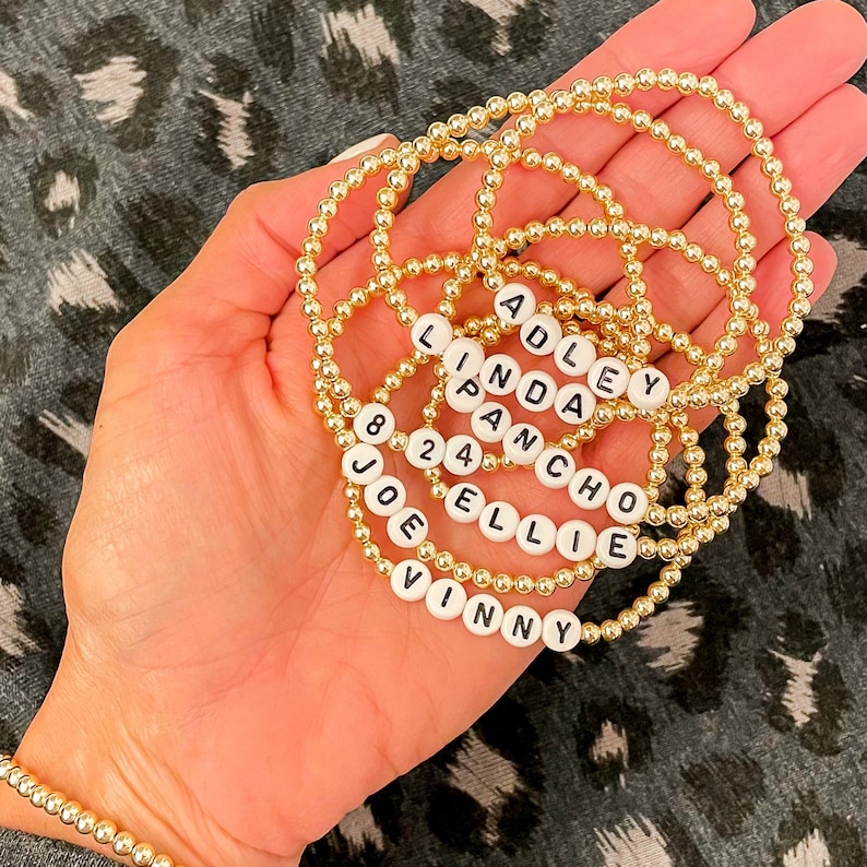 May include: A set of gold beaded bracelets with white beads spelling out the names "Adley", "Linda", "Pancho", "Ellie", "Joe", "Vinny", and "824".