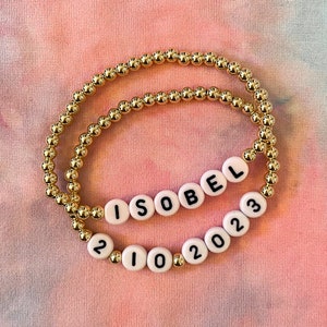 May include: Two gold beaded bracelets with white beads spelling out "ISOBEL" and "2102023".
