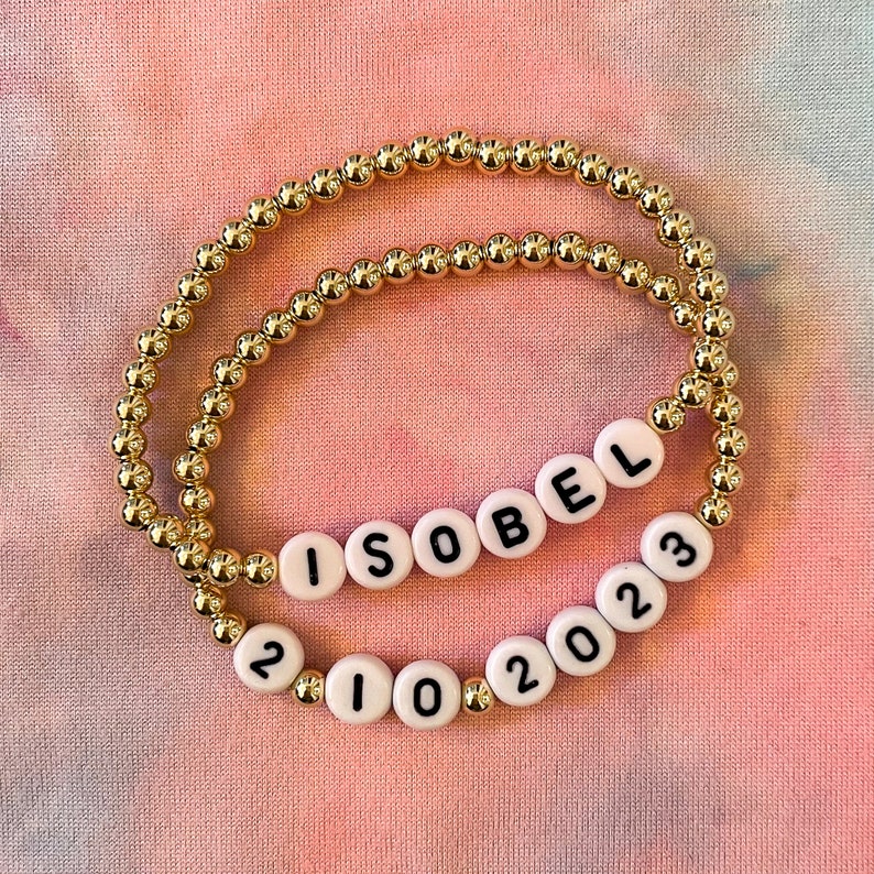 May include: Two gold beaded bracelets with white beads spelling out "ISOBEL" and "2102023".