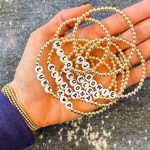 May include: A collection of gold bead bracelets with white beads spelling out names, including "Luke", "Ellie", "Mia", "Lost", "Edona", "Jell", "Bowie", and "MJ".