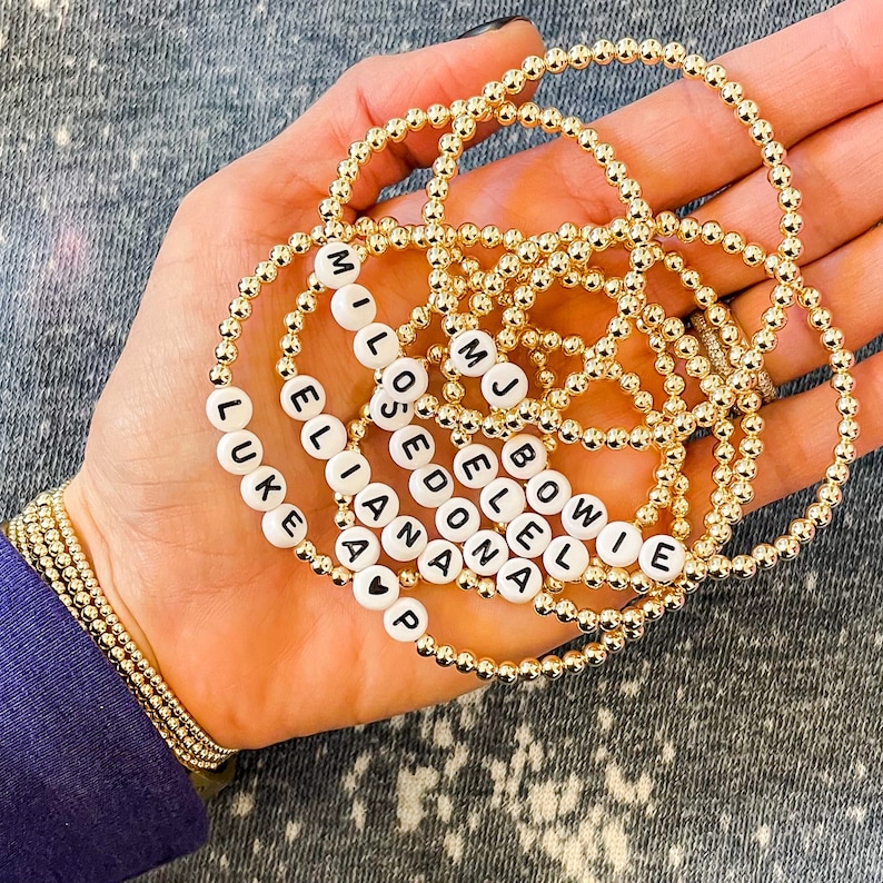 May include: A collection of gold bead bracelets with white beads spelling out names, including "Luke", "Ellie", "Mia", "Lost", "Edona", "Jell", "Bowie", and "MJ".
