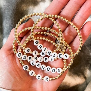 May include: Five gold beaded bracelets with white letter beads spelling out the names "Nick", "Landon", "Gabriel", "Brian", and "Brinley".