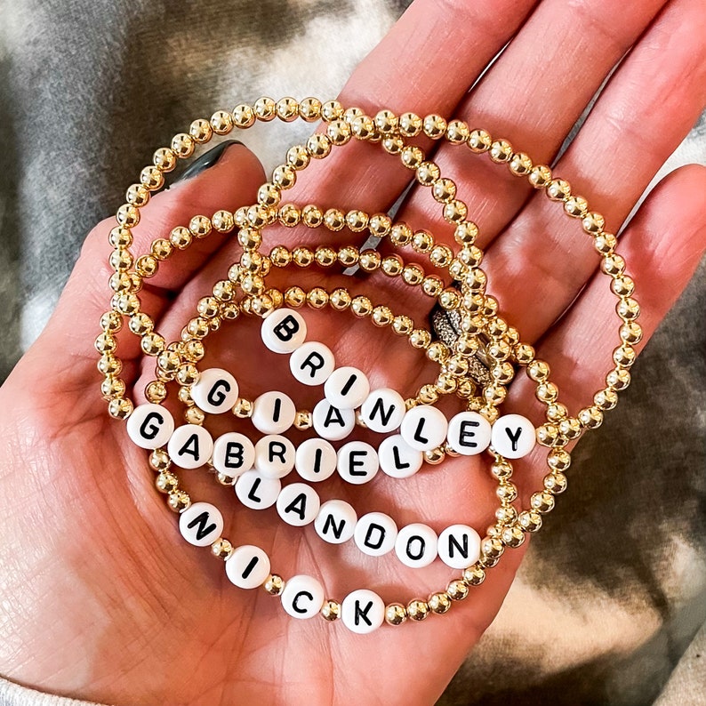 May include: Five gold beaded bracelets with white letter beads spelling out the names "Nick", "Landon", "Gabriel", "Brian", and "Brinley".