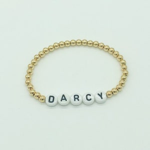 May include: Gold bead bracelet with the name "DARCY" spelled out in white beads with black lettering.