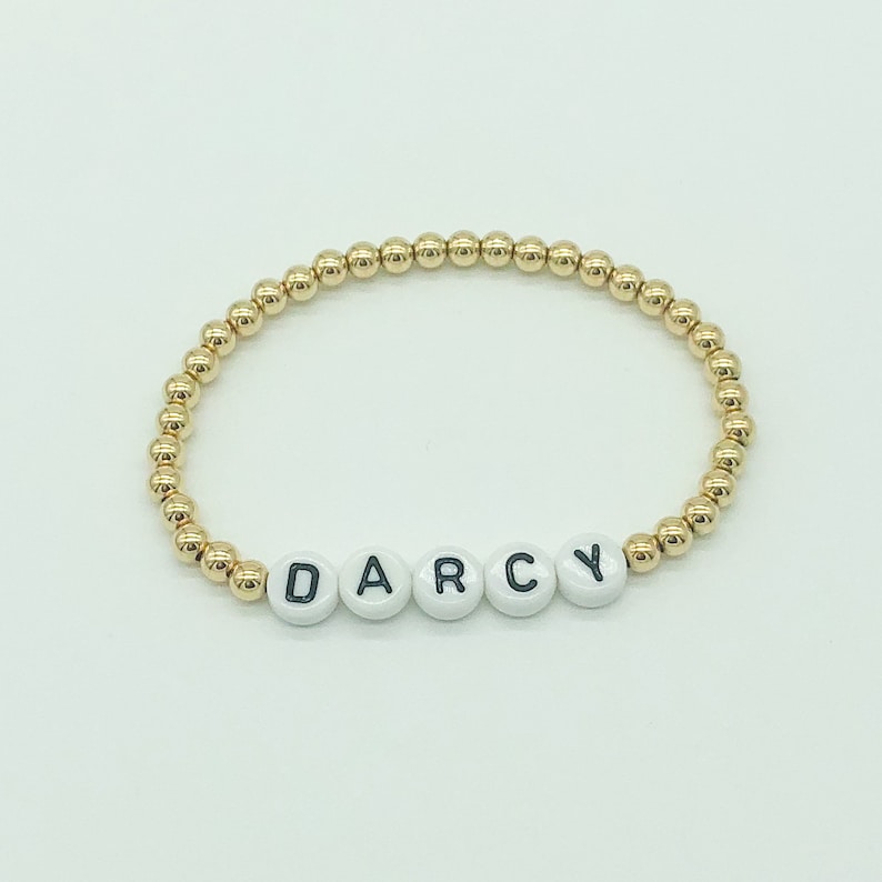 May include: Gold bead bracelet with the name "DARCY" spelled out in white beads with black lettering.