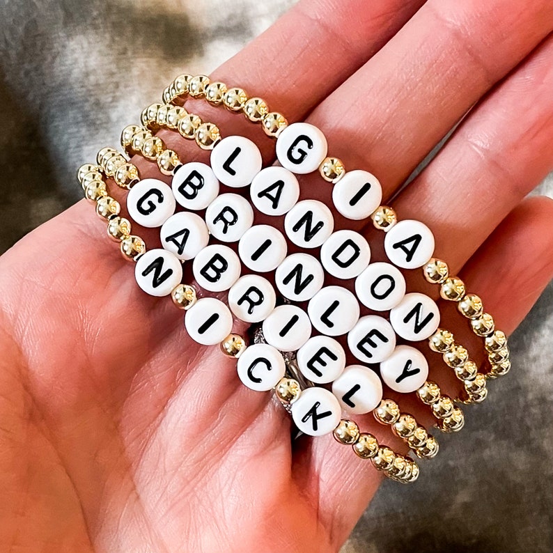 May include: Five gold bead bracelets with white letter beads spelling out the names Landon, Gianna, Brinley, Gabby, and Nick.
