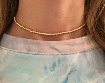 May include: A gold beaded choker necklace on a person wearing a light blue tie-dye shirt.