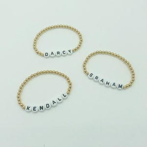 May include: Three gold bead bracelets with white letter beads spelling out the names "Darcy", "Graham", and "Kendall".