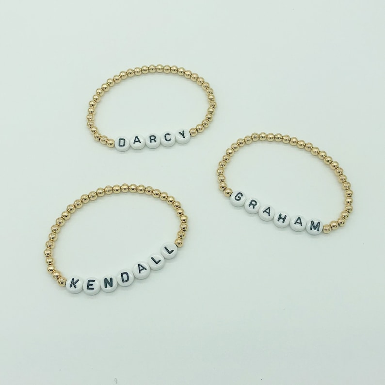 May include: Three gold bead bracelets with white letter beads spelling out the names "Darcy", "Graham", and "Kendall".