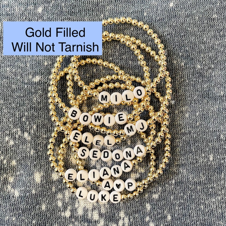 May include: Gold filled bracelets with white beads and black lettering spelling out names like "MILO", "BOWIE", "EL", "SEDONA", "ELIANA", and "LUKE".