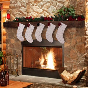 May include: Six white Christmas stockings with black polka dots hang above a fireplace. Each stocking has a different colored pom-pom. The fireplace is made of stone and has a wooden mantel decorated with greenery and ornaments.