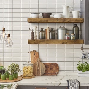 May include: Two wooden shelves mounted on a white tiled wall in a kitchen. The shelves are filled with various kitchen items, including jars, bowls, and a metal pot. There are also three hanging light fixtures with cage-style shades.