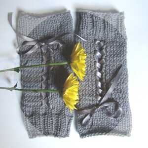 Puede incluir: Un par de calentadores de piernas grises tejidos con detalles de cinta y lazos. Los calentadores de piernas est&aacute;n decorados con una cinta gris que sube por la parte delantera y se ata con un lazo. Dos flores amarillas se colocan delante de los calentadores.