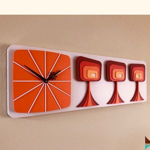 May include: A mid-century modern wall clock with a white background and orange clock face. The clock has three stylized trees with orange, red, and brown accents.