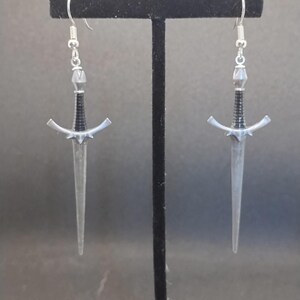May include: A pair of silver sword earrings with black accents. The swords have a pointed tip and a decorative handle.
