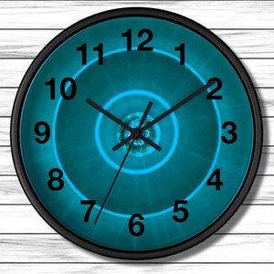 May include: Round wall clock with a black frame and a teal face. The clock face features a concentric spiral design with black numerals and hands. The background is a white wood grain.