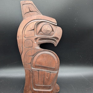 Può includere: Scultura in legno intagliato di un uccello, probabilmente un'aquila, in stile tradizionale. La scultura è di colore marrone scuro e presenta intagli dettagliati delle caratteristiche dell'uccello. L'opera d'arte è esposta su uno sfondo scuro.