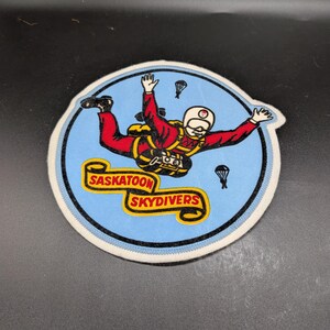 May include: A circular patch featuring a skydiver in a red jumpsuit and white helmet against a light blue background. The patch has the words "Saskatoon Skydivers" in yellow. Two small parachutes are also visible.