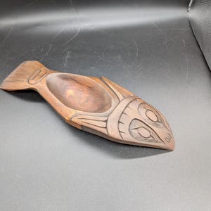 Puede incluir: Un cuenco de madera tallado a mano con forma de pez. El cuenco es marrón con detalles tallados que representan las características del pez. El cuenco tiene una hendidura profunda de forma ovalada en el centro.